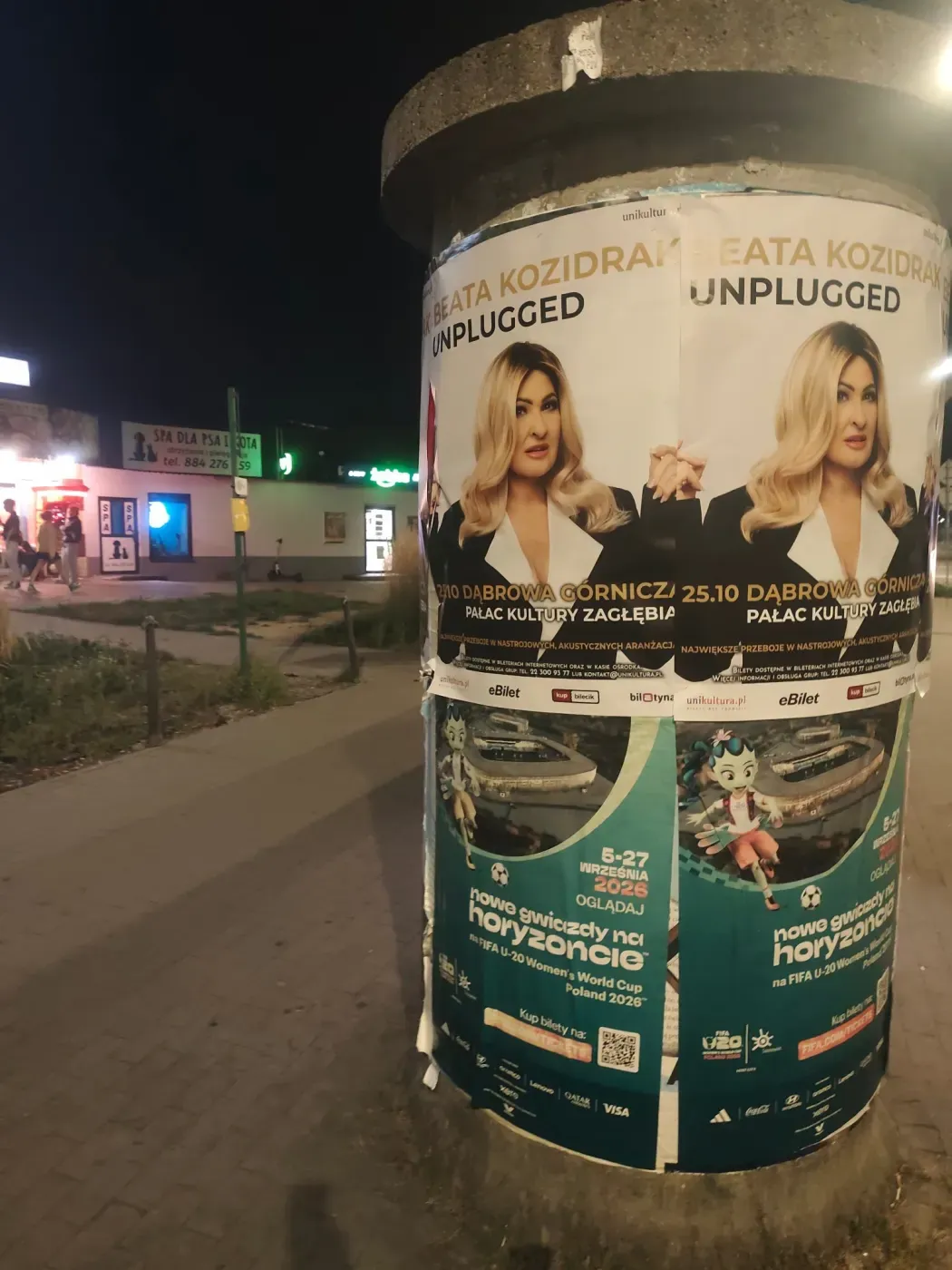 Okrągły słup ogłoszeniowy z plakatami Beata Kozidrak Unplugged przy chodniku