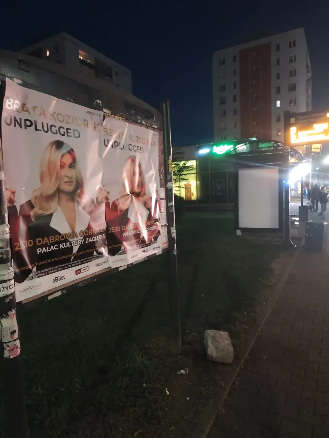 Plakaty koncertu na tablicy ogłoszeniowej obok przystanku i chodnika