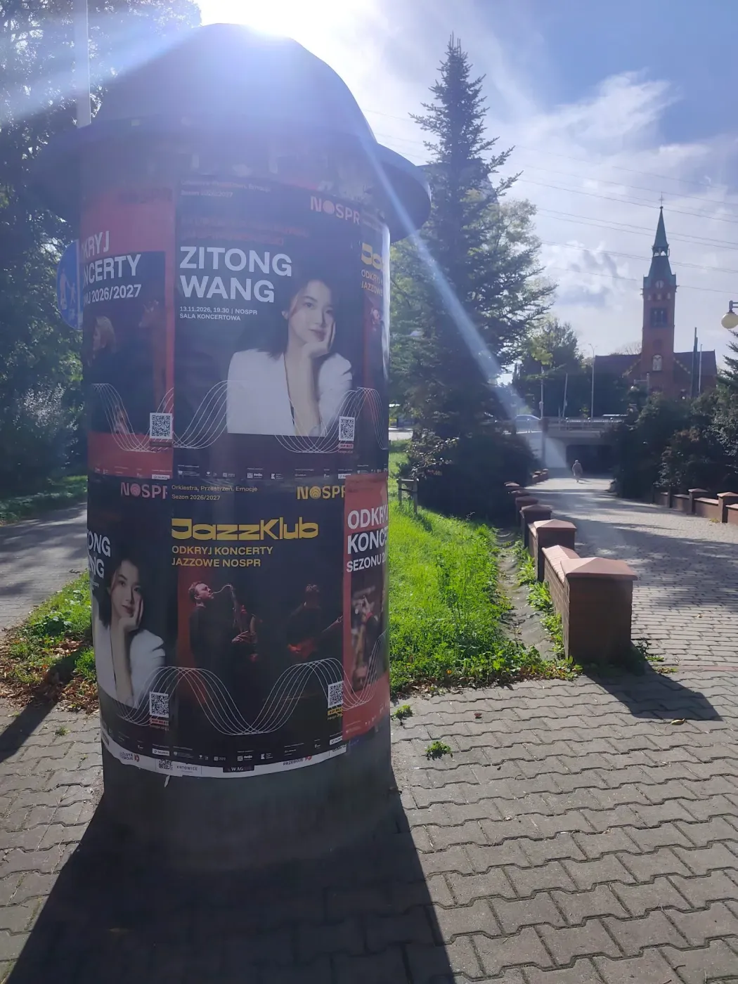Słup z plakatami koncertowymi przy zielonym skwerze i kościele w Katowicach