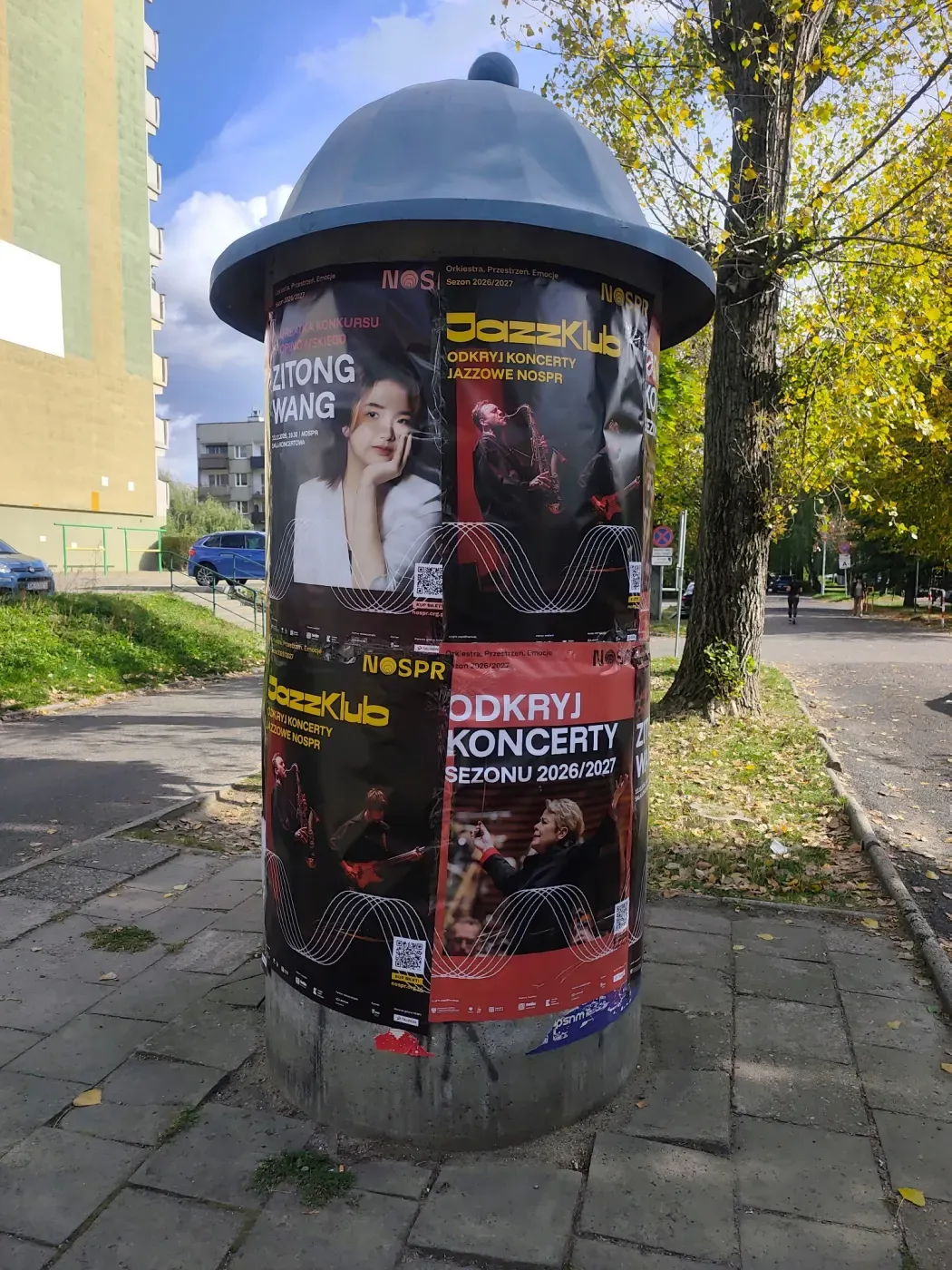 Okrągły słup ogłoszeniowy z plakatami koncertowymi przy osiedlowym chodniku w Katowicach