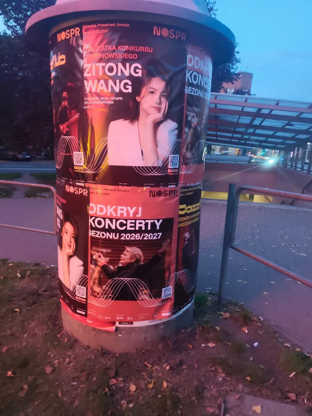 Plakaty koncertowe na słupie ogłoszeniowym obok wiaty przystankowej i chodnika