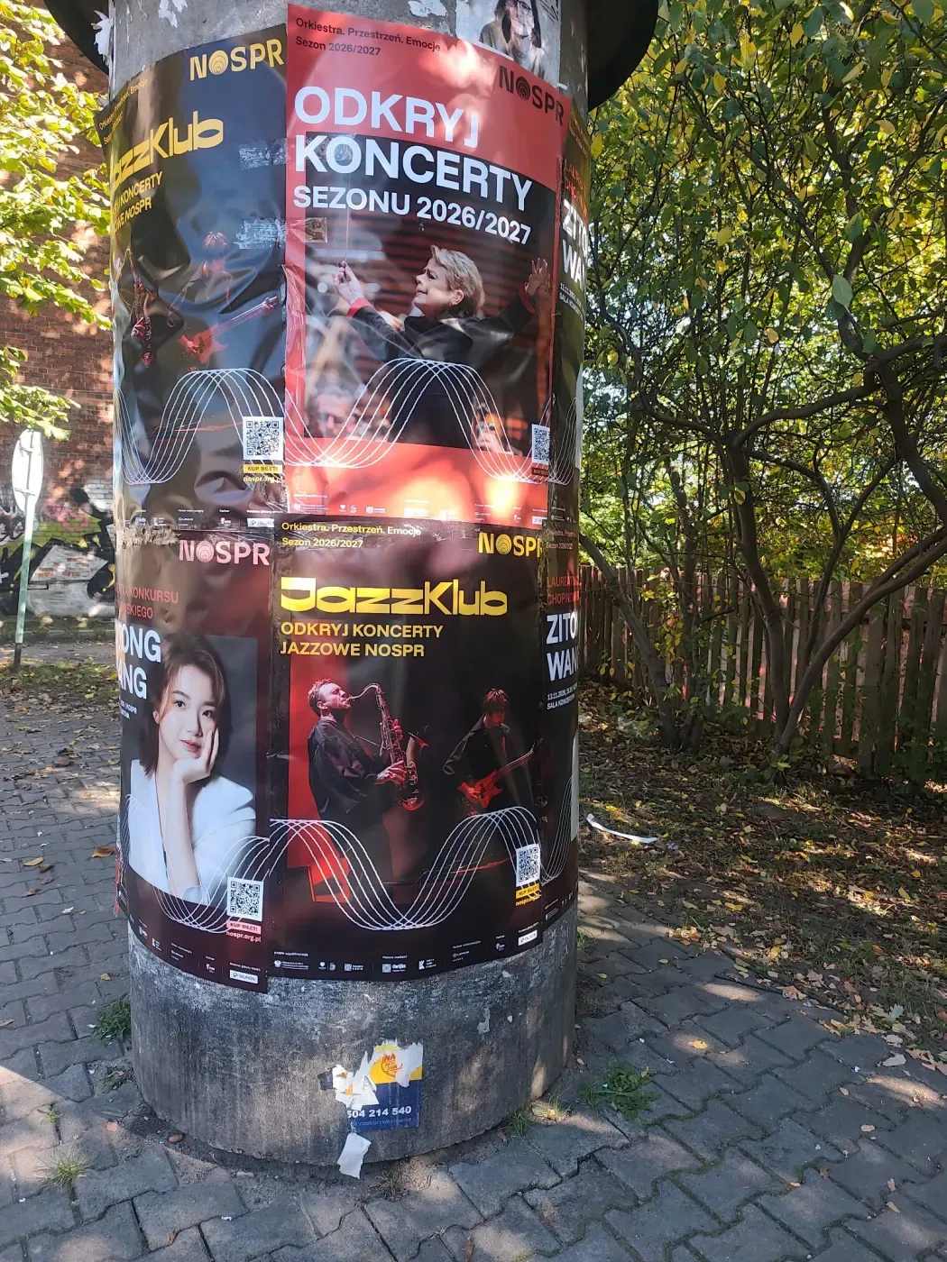 Czerwono-czarne plakaty sezonu koncertowego i JazzKlubu na słupie ogłoszeniowym wśród zieleni