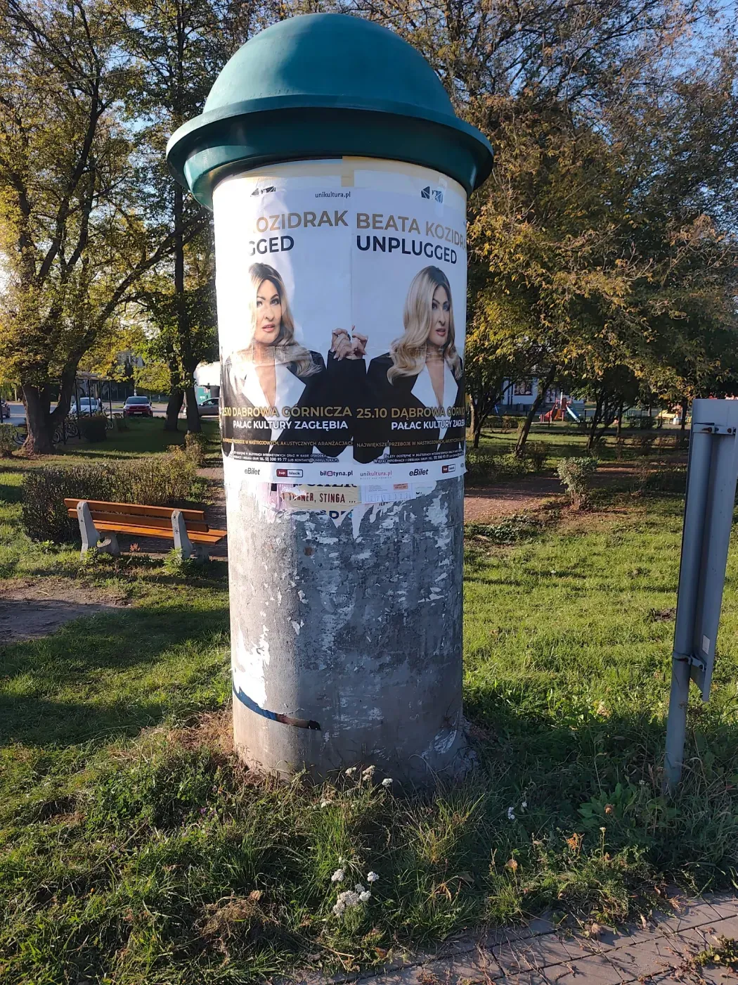 Plakaty Beata Kozidrak Unplugged na okrągłym słupie ogłoszeniowym w zielonej części miasta