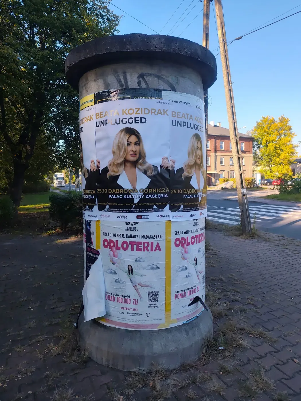 Słup ogłoszeniowy z plakatami Beata Kozidrak Unplugged przy chodniku i przejściu dla pieszych