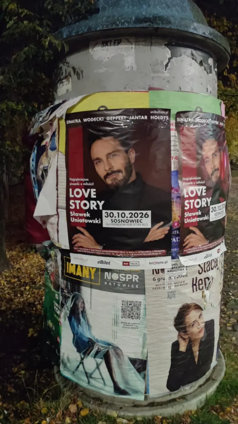 Dwa plakaty Love Story na okrągłym słupie ogłoszeniowym wśród innych afiszy.