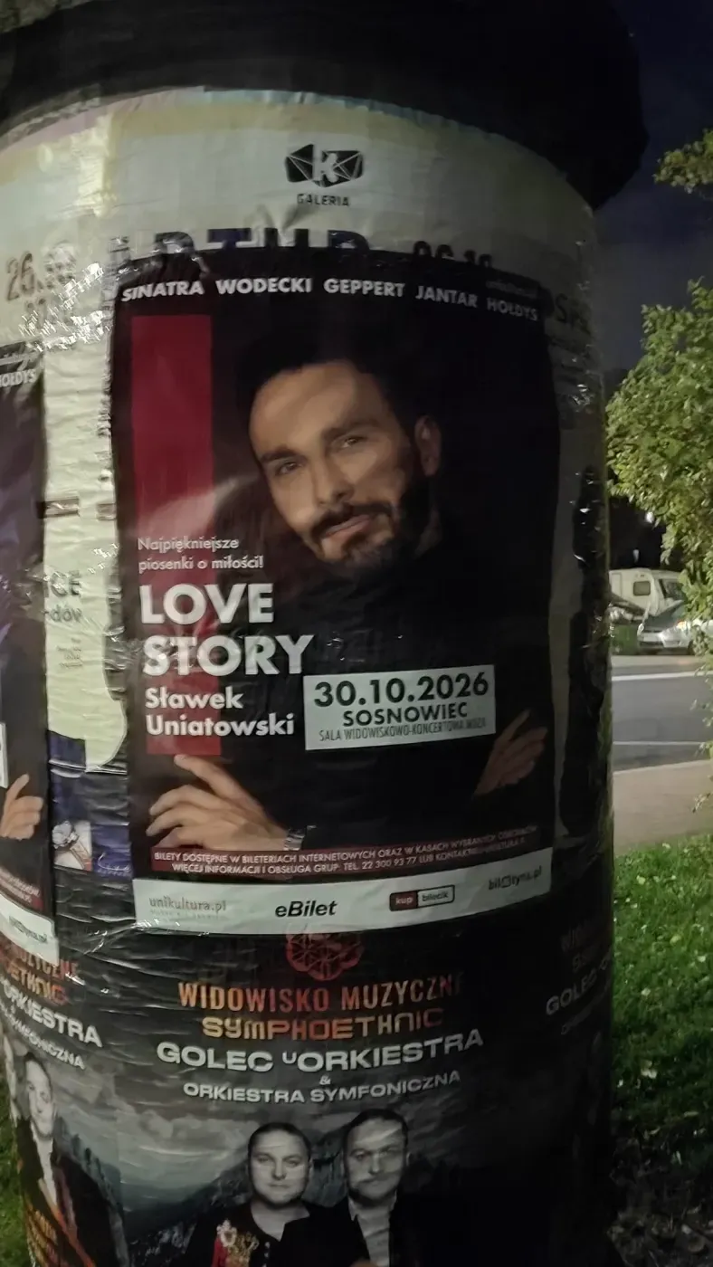 Plakat Love Story Sławka Uniatowskiego na dużym słupie przy ulicy i zieleni.